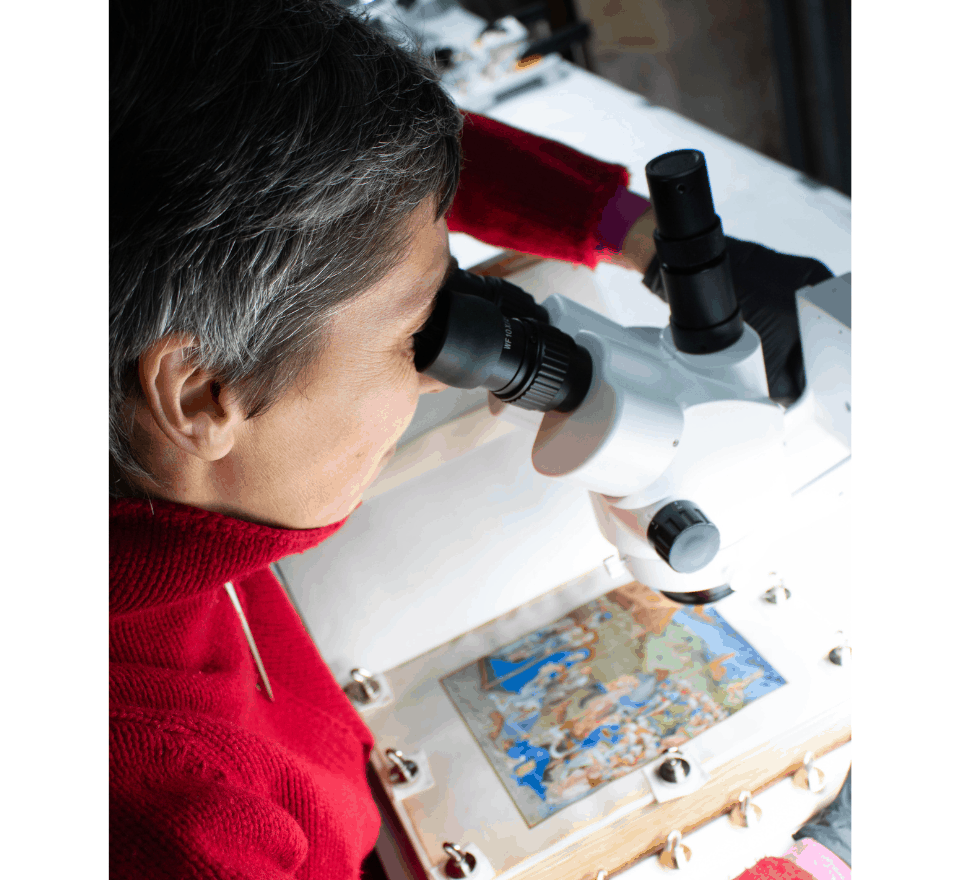 The restorer uses a binocular microscope to apply a tiny drop of neutral adhesive with a fine brush, under a piece of the paint layer that is peeling away from the parchment. © Château de Chantilly